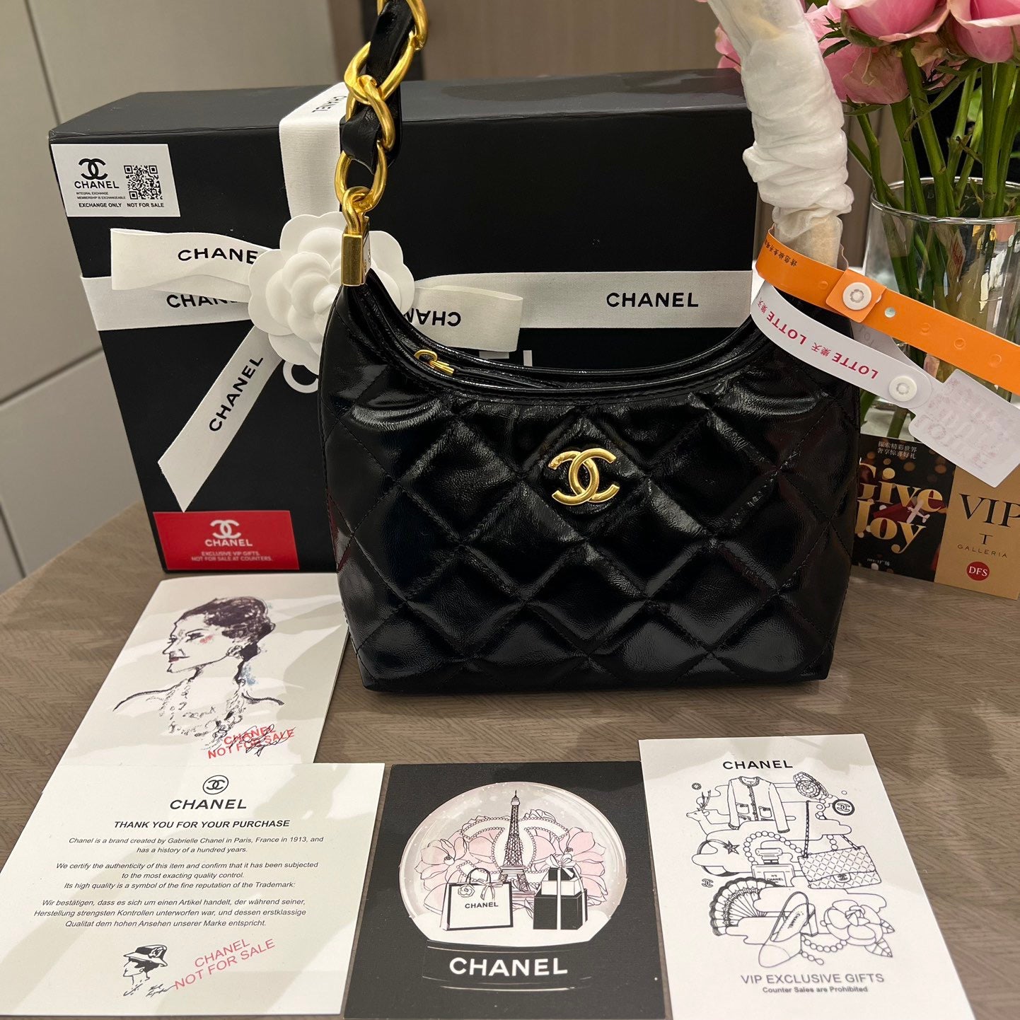 Black quilted handbag with gold chain and hardware, accompanied by Chanel packaging and documents on a wooden surface.